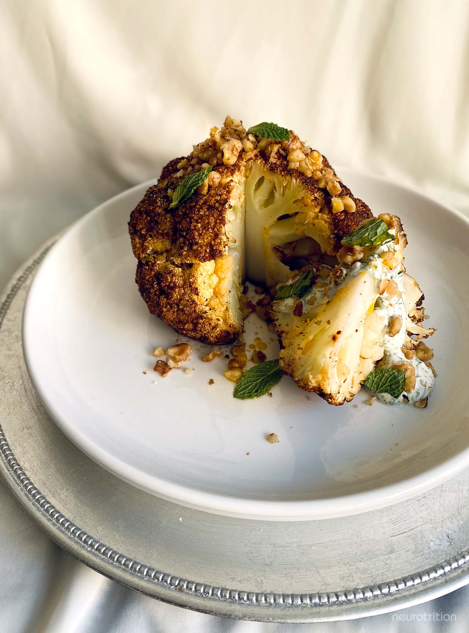 Baharat Whole Roasted Cauliflower w/ Garlic Yogurt & Walnuts Whole roasted cauliflower with wedge cut out topped with herbs and yogurt sauce on a white plate
