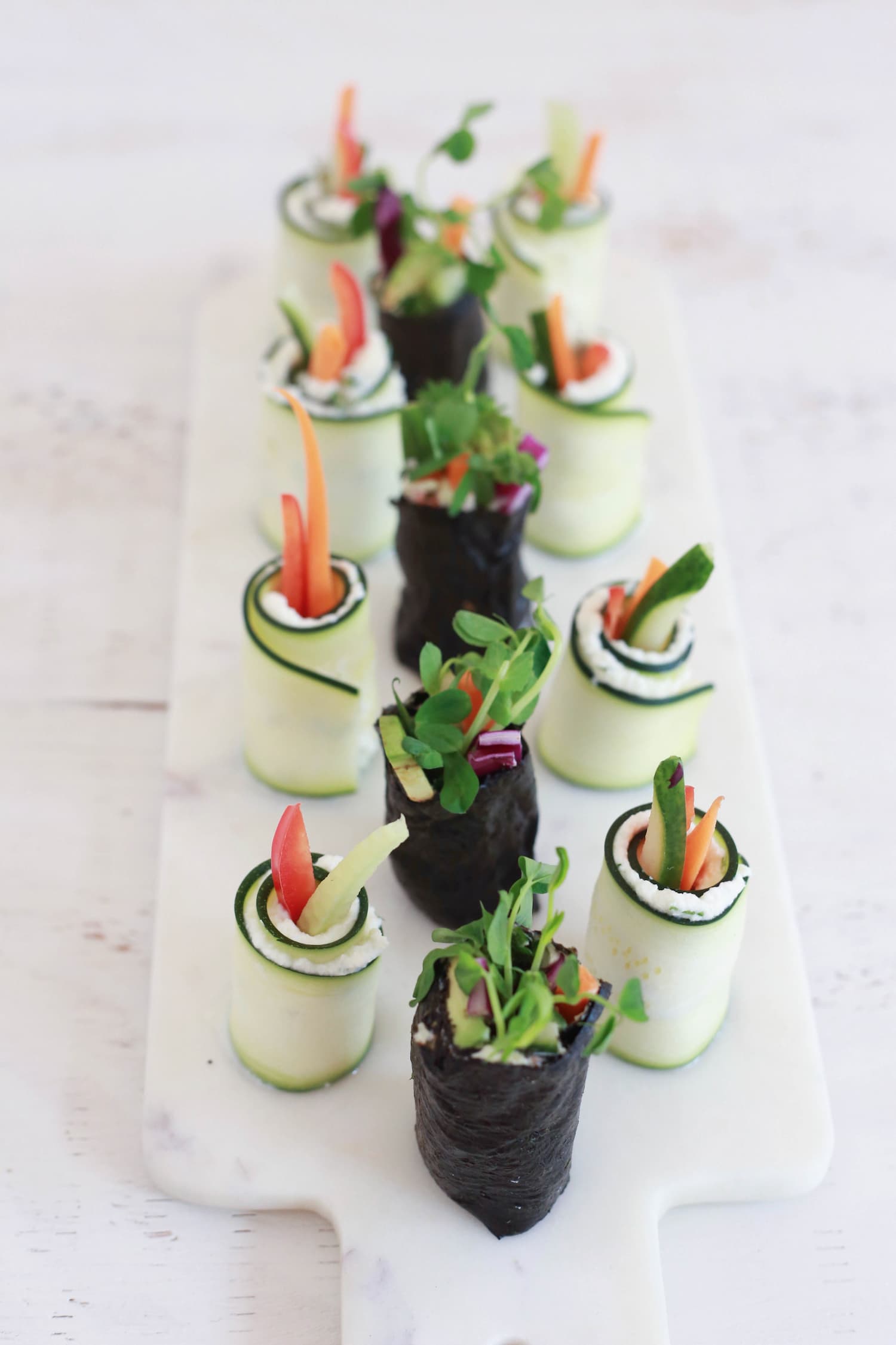 Rice-Free Veggie Sushi Veggie sushi wrapped in nori and raw zuchinni, on a white marble board.
